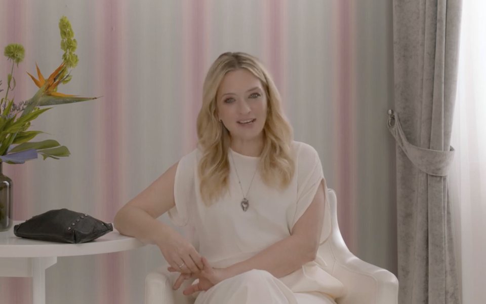 Conduttrice bionda seduta su sfondo a righe rosa per il branded content My Beauty Bag di ELLE